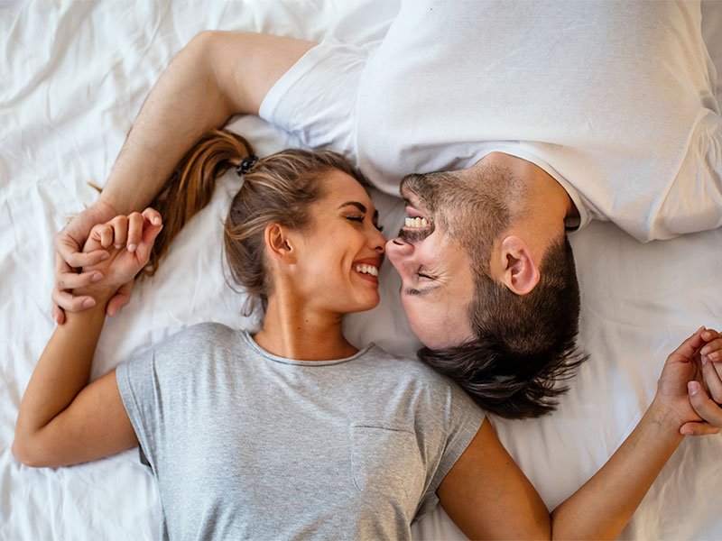 Life Extension, smiling, joyful woman and man in white and grey T-shirts on their backs on the bed holding hands